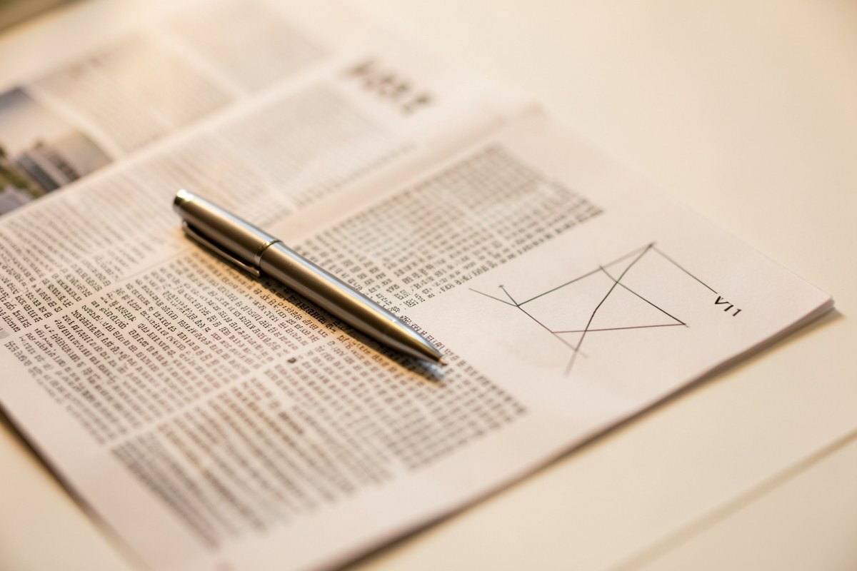 A close-up of a newspaper financial section with a pen pointing at a small line chart.